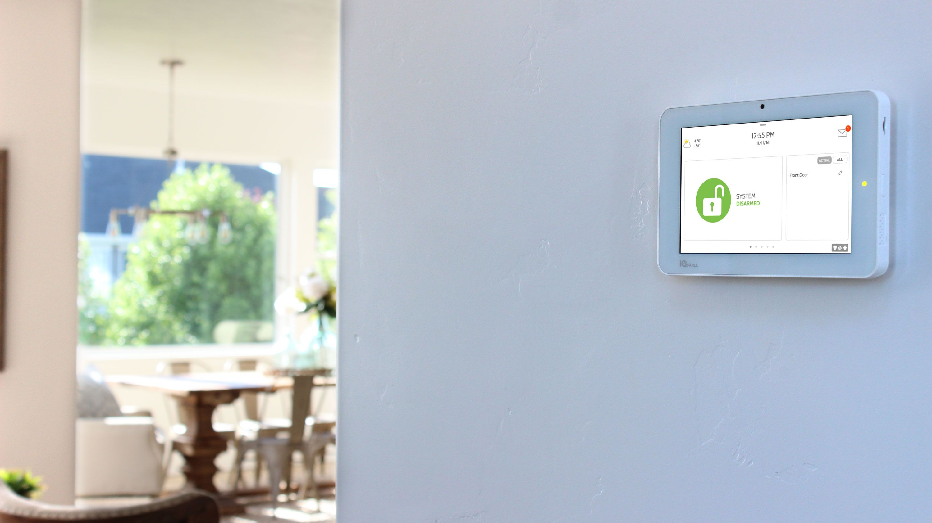 A home security touchscreen panel mounted on a beige wall near the front door, displaying a user interface.