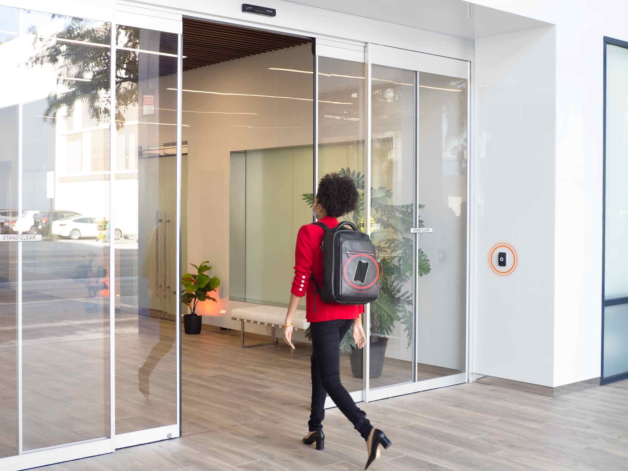 Woman wearing a red blazer and backpack entering a modern building through automatic glass doors.