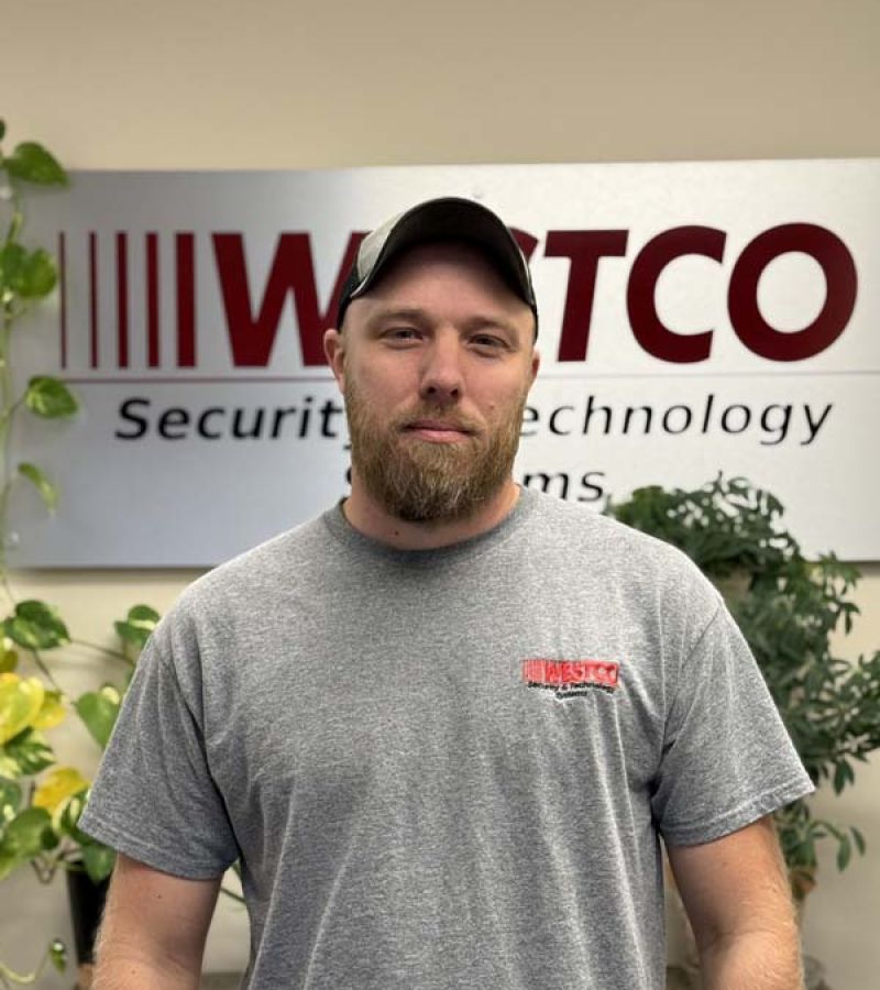 A bearded man wearing a gray Westco T-shirt and a baseball cap stands in front of a “WESTCO Security & Technology Systems” sign, with green potted plants in the background.
