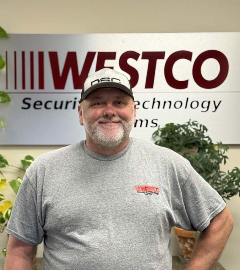 A smiling man wearing a gray Westco T-shirt and a black-and-white DSC baseball cap stands in front of a “WESTCO Security & Technology Systems” sign, with green potted plants nearby.