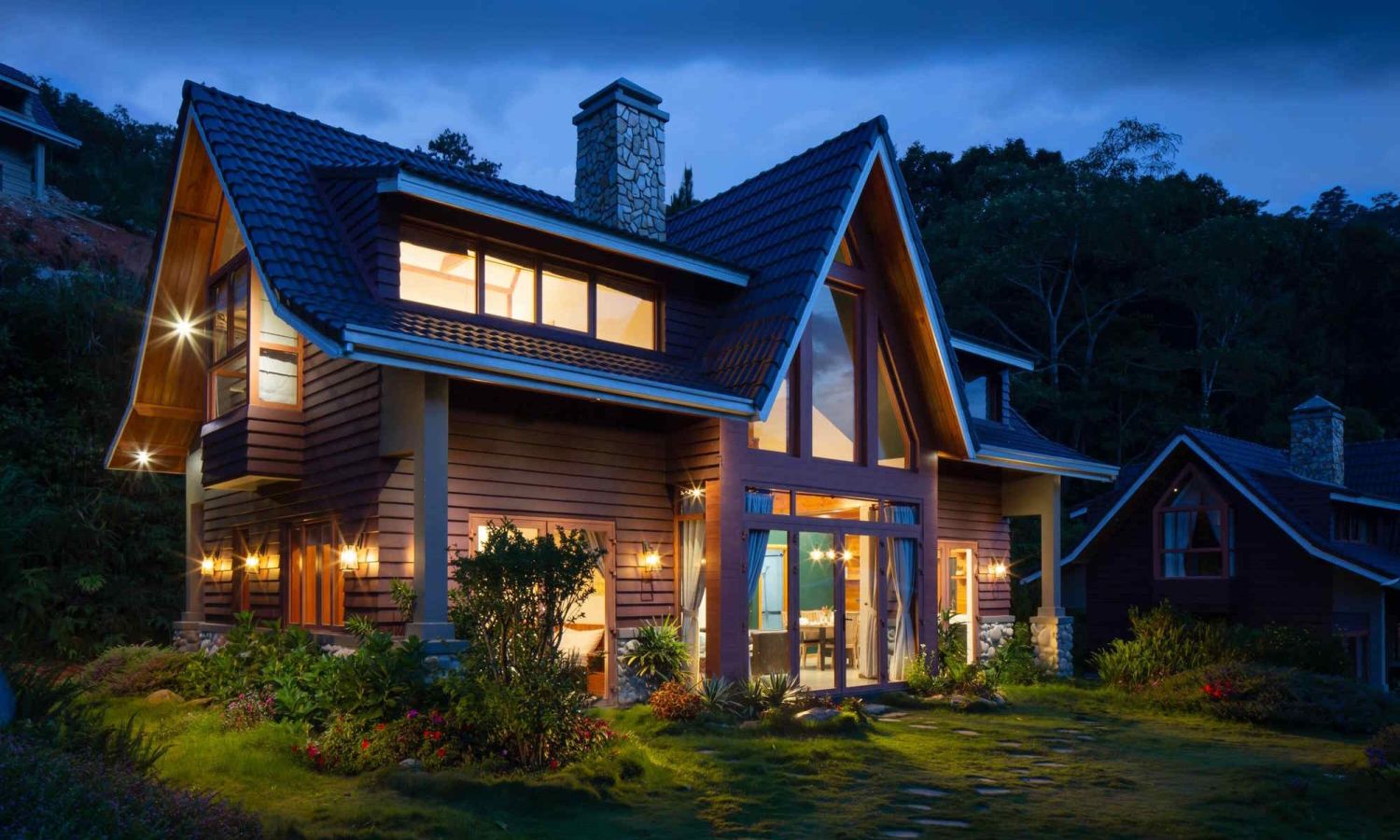 A modern craftsman-style home at dusk with large windows, steep rooflines, and warm exterior lighting illuminating the landscaped yard and surrounding greenery.