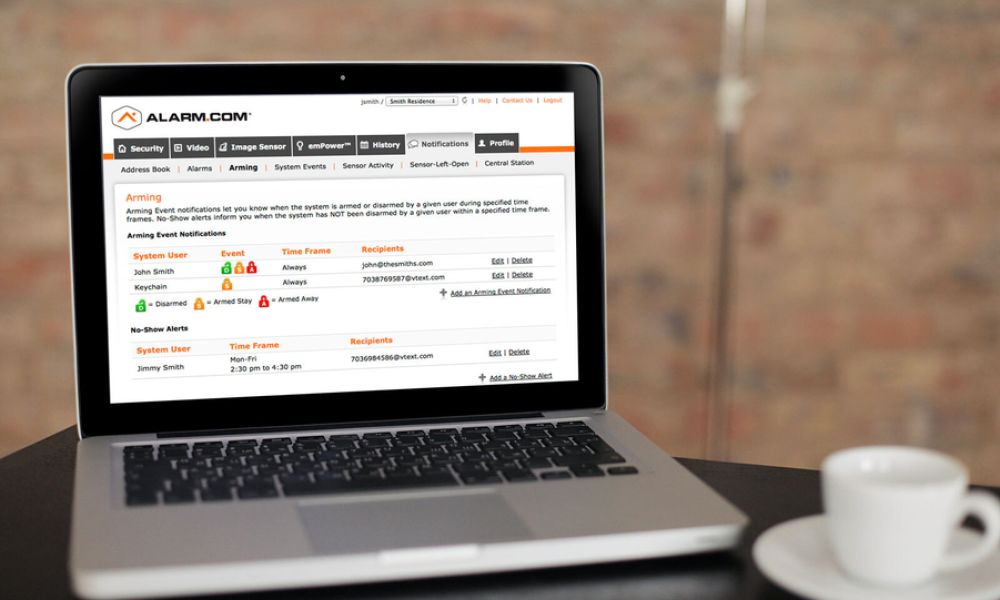 Open laptop on a table displaying the Alarm.com web dashboard with arming notifications, next to a white coffee cup.