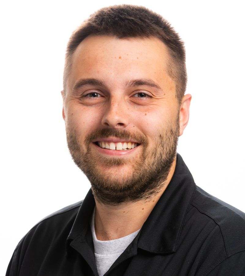 A smiling man in glasses wearing a red “WESTCO” polo shirt stands in front of a “WESTCO Security and Technology Systems” sign, with green potted plants behind him.