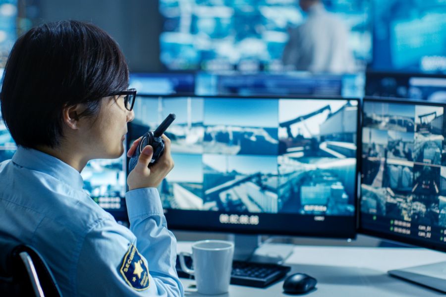 Security operator with a radio reviews camera feeds of an industrial yard on multiple monitors in a monitoring center.