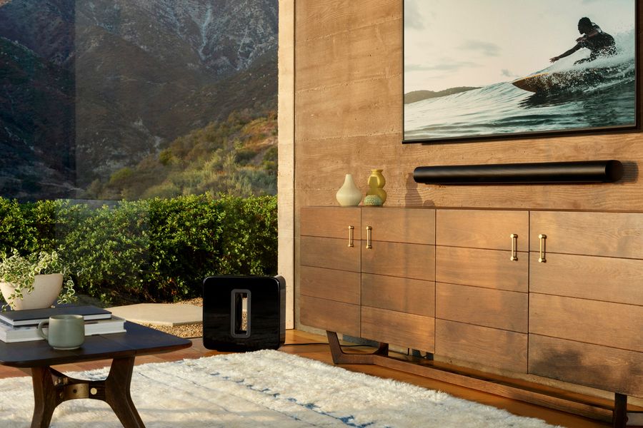 Black Sonos Arc soundbar mounted below a wall-mounted TV, with a Sonos Sub subwoofer on the floor nearby.