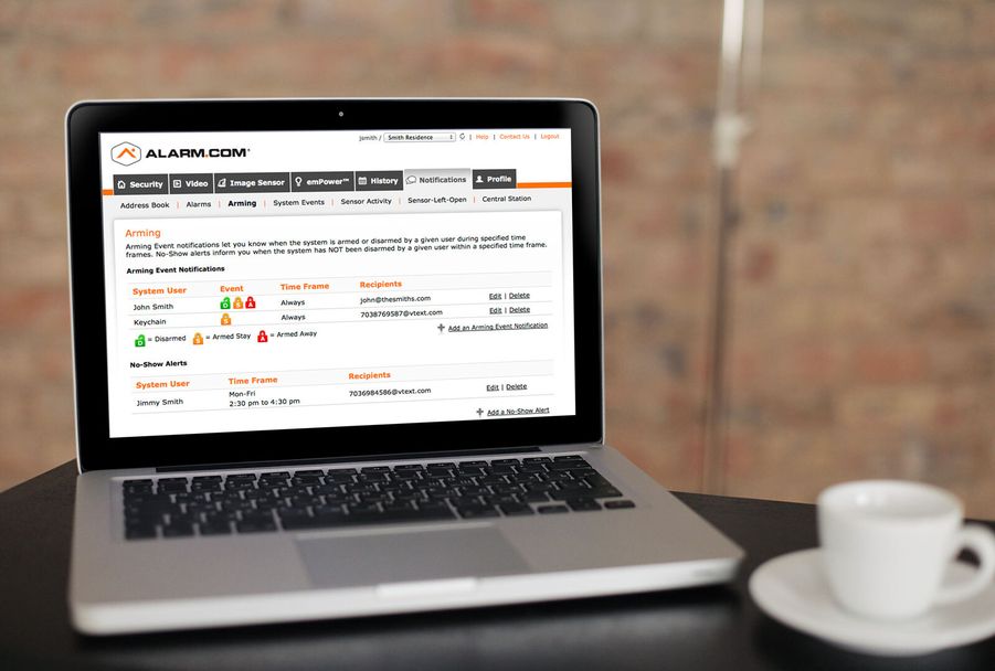 Open laptop on a table displaying the Alarm.com web dashboard with arming notifications, next to a white coffee cup.