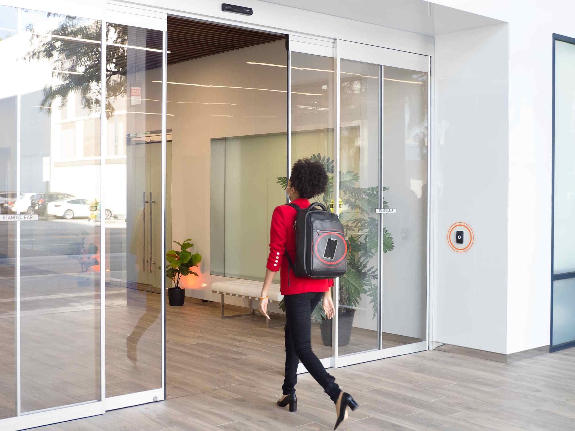 Woman wearing a red blazer and backpack entering a modern building through automatic glass doors.