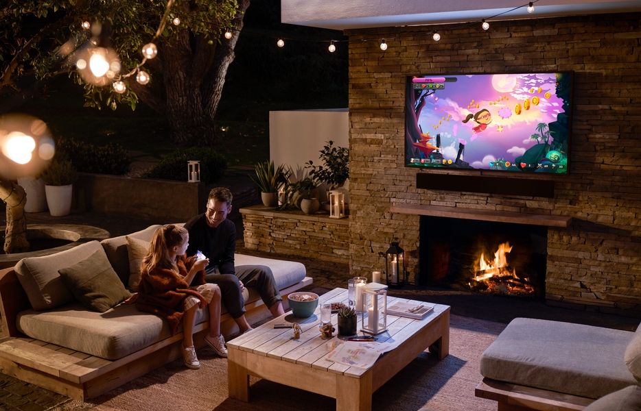 A man and young girl sit on an outdoor patio watching a video game on a wall-mounted TV above a fireplace.
