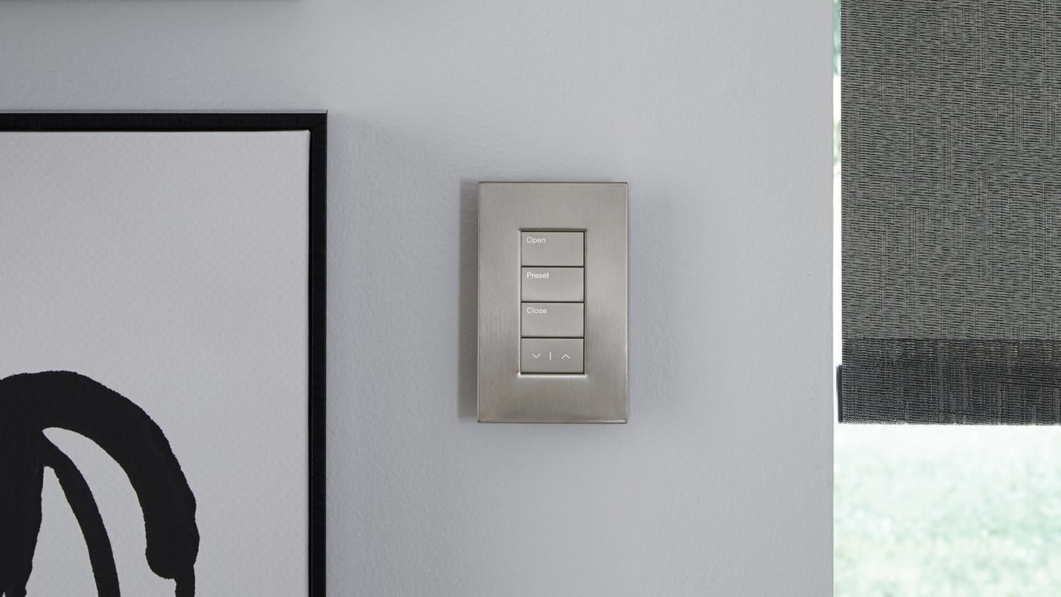 Sleek silver Lutron wall switch with buttons labeled "Open," "Preset," and "Close," next to a window with a grey shade.